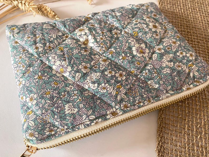 May include: A quilted floral clutch in shades of teal, white, and yellow. The rectangular pouch features a repeating flower pattern and a gold-toned zipper closure. The fabric has a diamond-shaped quilted design.