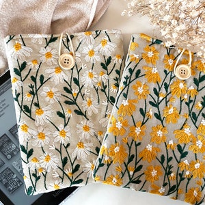 Könnte beinhalten: Zwei beigefarbene E-Reader-Hüllen aus Leinen mit gestickten Blumenmustern. Eine Hülle zeigt weiße Blumen mit gelben Zentren, die andere zeigt gelbe Blumen mit weißen Zentren. Beide Hüllen haben einen Holzknopf.