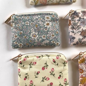 May include: Several small, rectangular zippered pouches with floral patterns are arranged on a white surface. The pouches feature various designs, including white flowers on a teal background, and pink roses on a cream background. Each pouch has a gold-colored zipper.