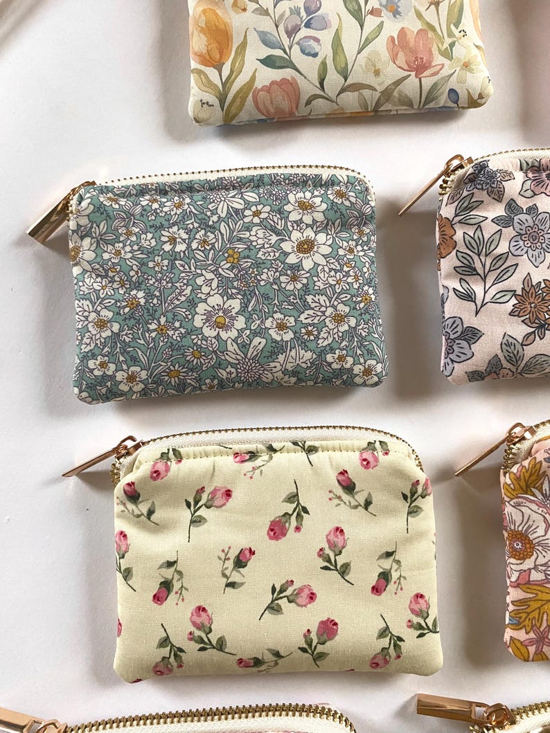 May include: Several small, rectangular zippered pouches with floral patterns are arranged on a white surface. The pouches feature various designs, including white flowers on a teal background, and pink roses on a cream background. Each pouch has a gold-colored zipper.