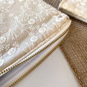 May include: Close-up of a beige clutch with intricate white floral embroidery. The clutch features a gold zipper and is partially open, revealing the interior. A smaller matching item is visible in the background.