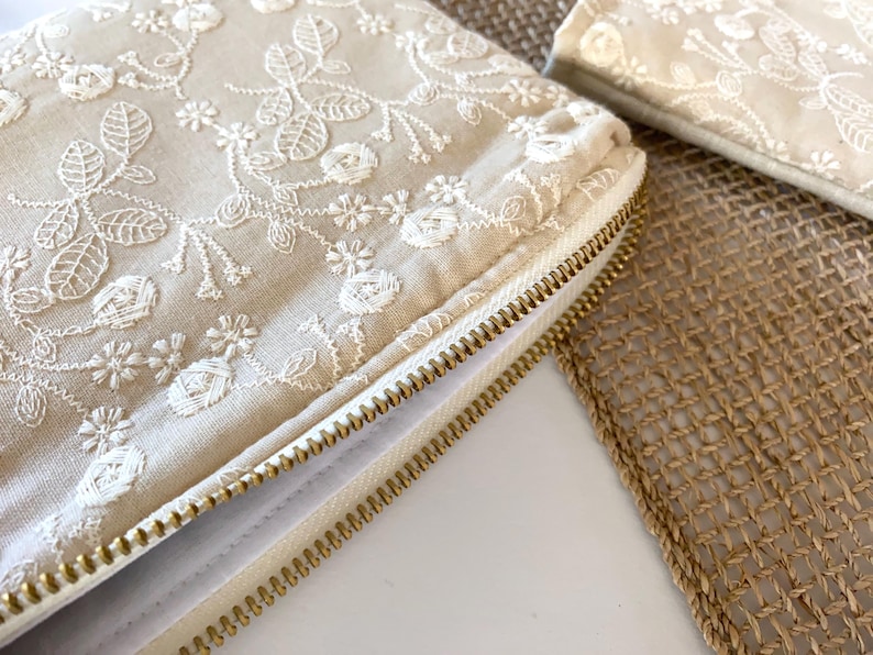 May include: Close-up of a beige clutch with intricate white floral embroidery. The clutch features a gold zipper and is partially open, revealing the interior. A smaller matching item is visible in the background.