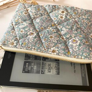 May include: A floral-patterned, quilted pouch in shades of blue, white, and yellow, with a gold zipper. The pouch is open, revealing an e-reader with book covers visible on the screen. The pouch is designed to protect the e-reader.
