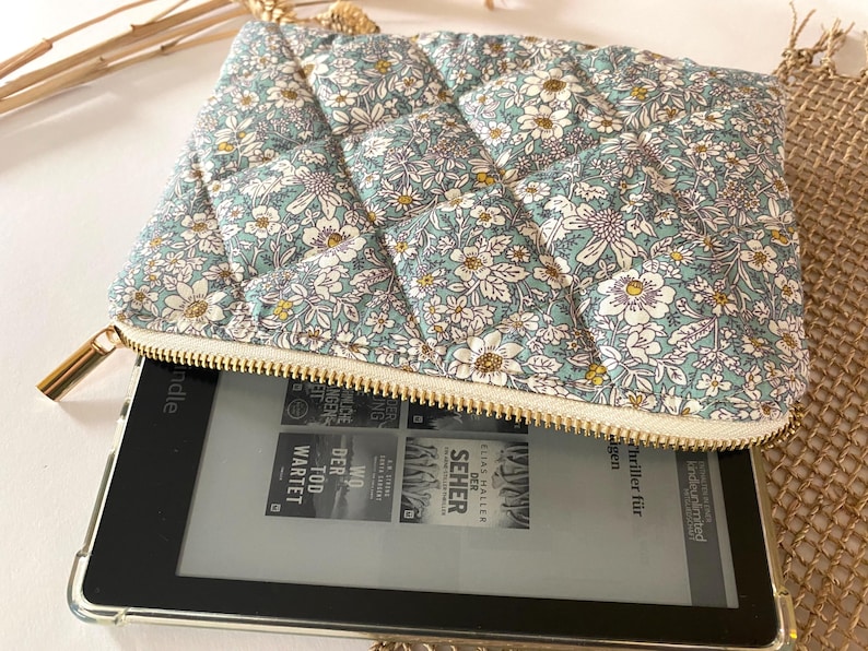 May include: A floral-patterned, quilted pouch in shades of blue, white, and yellow, with a gold zipper. The pouch is open, revealing an e-reader with book covers visible on the screen. The pouch is designed to protect the e-reader.