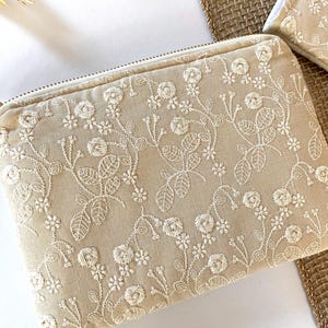 May include: Beige zippered pouch with intricate white floral embroidery. The design features detailed roses, leaves, and vines, creating a delicate and elegant pattern. The pouch has a gold-colored zipper pull and is likely used for cosmetics or small items.