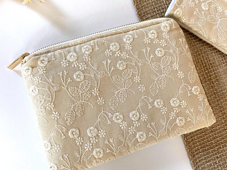 May include: Beige zippered pouch with intricate white floral embroidery. The design features detailed roses, leaves, and vines, creating a delicate and elegant pattern. The pouch has a gold-colored zipper pull and is likely used for cosmetics or small items.