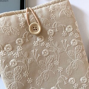 Pu&ograve; includere: Una custodia per libri in lino beige con un ricamo floreale bianco e una chiusura a bottone beige.