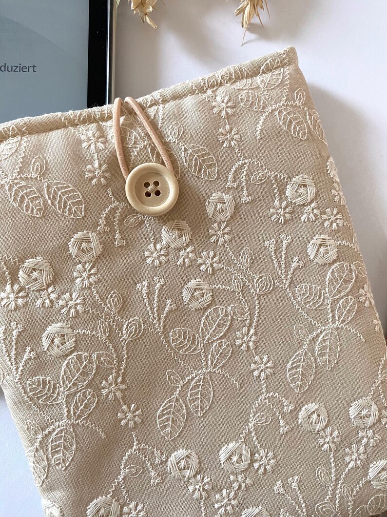 Pu&ograve; includere: Una custodia per libri in lino beige con un ricamo floreale bianco e una chiusura a bottone beige.