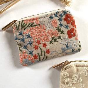K&ouml;nnte beinhalten: Beige bestickte Tasche mit goldenem Rei&szlig;verschluss. Die Tasche hat ein Blumenmuster mit korallenfarbenen, blauen und gr&uuml;nen F&auml;den. Die Stickerei umfasst Blumen und Bl&auml;tter. Eine zweite Tasche mit wei&szlig;em Blumenmuster ist sichtbar.