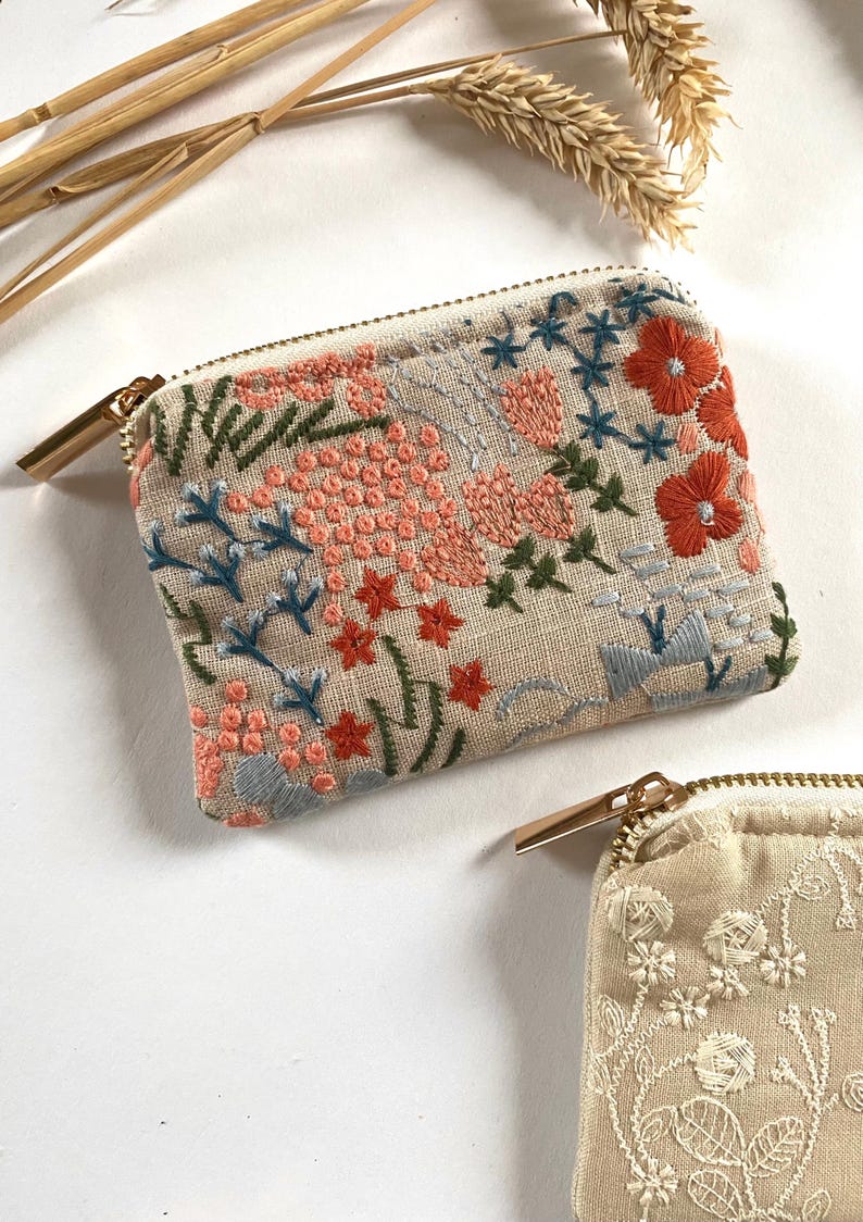 K&ouml;nnte beinhalten: Beige bestickte Tasche mit goldenem Rei&szlig;verschluss. Die Tasche hat ein Blumenmuster mit korallenfarbenen, blauen und gr&uuml;nen F&auml;den. Die Stickerei umfasst Blumen und Bl&auml;tter. Eine zweite Tasche mit wei&szlig;em Blumenmuster ist sichtbar.
