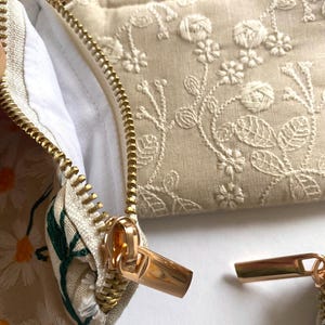 K&ouml;nnte beinhalten: Nahaufnahme von bestickten Kosmetiktaschen mit goldenen Rei&szlig;verschl&uuml;ssen. Eine Tasche hat ein G&auml;nsebl&uuml;mchenmuster, eine andere zeigt Blumenstickerei. Die Taschen sind beige und wei&szlig;, mit wei&szlig;em Innenfutter. Die Rei&szlig;verschl&uuml;sse sind goldfarben.