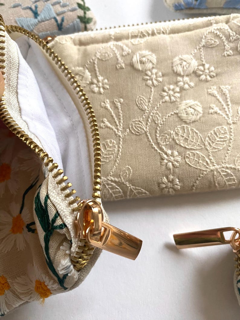 K&ouml;nnte beinhalten: Nahaufnahme von bestickten Kosmetiktaschen mit goldenen Rei&szlig;verschl&uuml;ssen. Eine Tasche hat ein G&auml;nsebl&uuml;mchenmuster, eine andere zeigt Blumenstickerei. Die Taschen sind beige und wei&szlig;, mit wei&szlig;em Innenfutter. Die Rei&szlig;verschl&uuml;sse sind goldfarben.