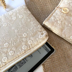 May include: Beige embroidered fabric clutch with a white zipper, and a matching case with a button closure. The floral embroidery is white. An e-reader is partially visible inside the clutch. The items are on a textured surface.