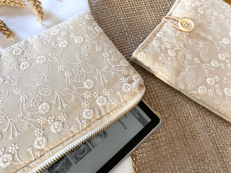 May include: Beige embroidered fabric clutch with a white zipper, and a matching case with a button closure. The floral embroidery is white. An e-reader is partially visible inside the clutch. The items are on a textured surface.