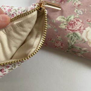 May include: Close-up of a floral print pouch with a gold zipper. The pouch is made of pink fabric with a pattern of roses and green leaves. The interior lining is a cream color.