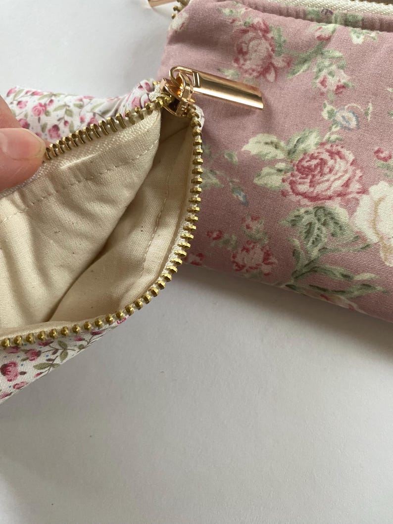 May include: Close-up of a floral print pouch with a gold zipper. The pouch is made of pink fabric with a pattern of roses and green leaves. The interior lining is a cream color.