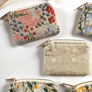 K&ouml;nnte beinhalten: Mehrere kleine, beige bestickte M&uuml;nzb&ouml;rsen mit floralen Designs. Die Geldb&ouml;rsen weisen Stickereien in verschiedenen Farben auf, darunter Rot, Rosa, Blau, Wei&szlig; und Gelb. Jede Geldb&ouml;rse hat einen goldenen Rei&szlig;verschluss und eine rechteckige Form.