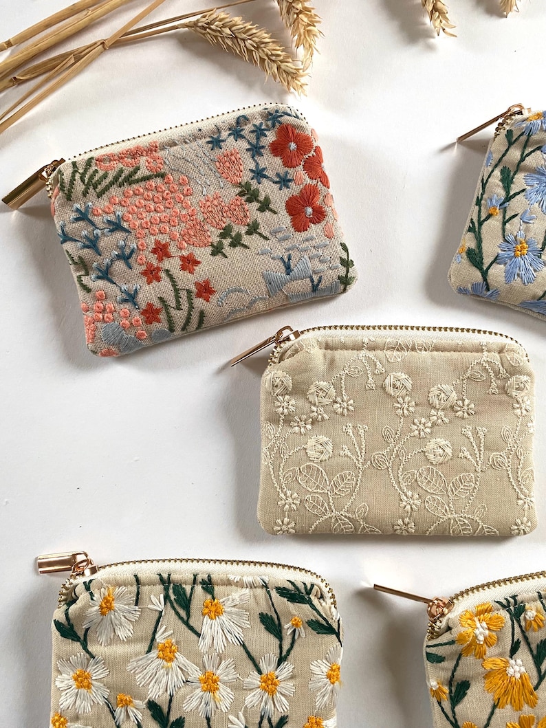 K&ouml;nnte beinhalten: Mehrere kleine, beige bestickte M&uuml;nzb&ouml;rsen mit floralen Designs. Die Geldb&ouml;rsen weisen Stickereien in verschiedenen Farben auf, darunter Rot, Rosa, Blau, Wei&szlig; und Gelb. Jede Geldb&ouml;rse hat einen goldenen Rei&szlig;verschluss und eine rechteckige Form.