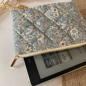 May include: A quilted floral print tablet case in shades of blue, white, and yellow, with a gold zipper. The case is open, revealing a tablet with text on the screen. The case is resting on a woven surface.
