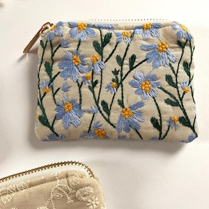 K&ouml;nnte beinhalten: Eine beige Rei&szlig;verschlusstasche mit gestickten blauen und gelben Blumen und gr&uuml;nen Stielen. Das florale Design bedeckt die Vorderseite der Tasche. Ein goldfarbener Rei&szlig;verschluss und Zipper sind sichtbar. Eine weitere Tasche mit floralem Design ist teilweise sichtbar.