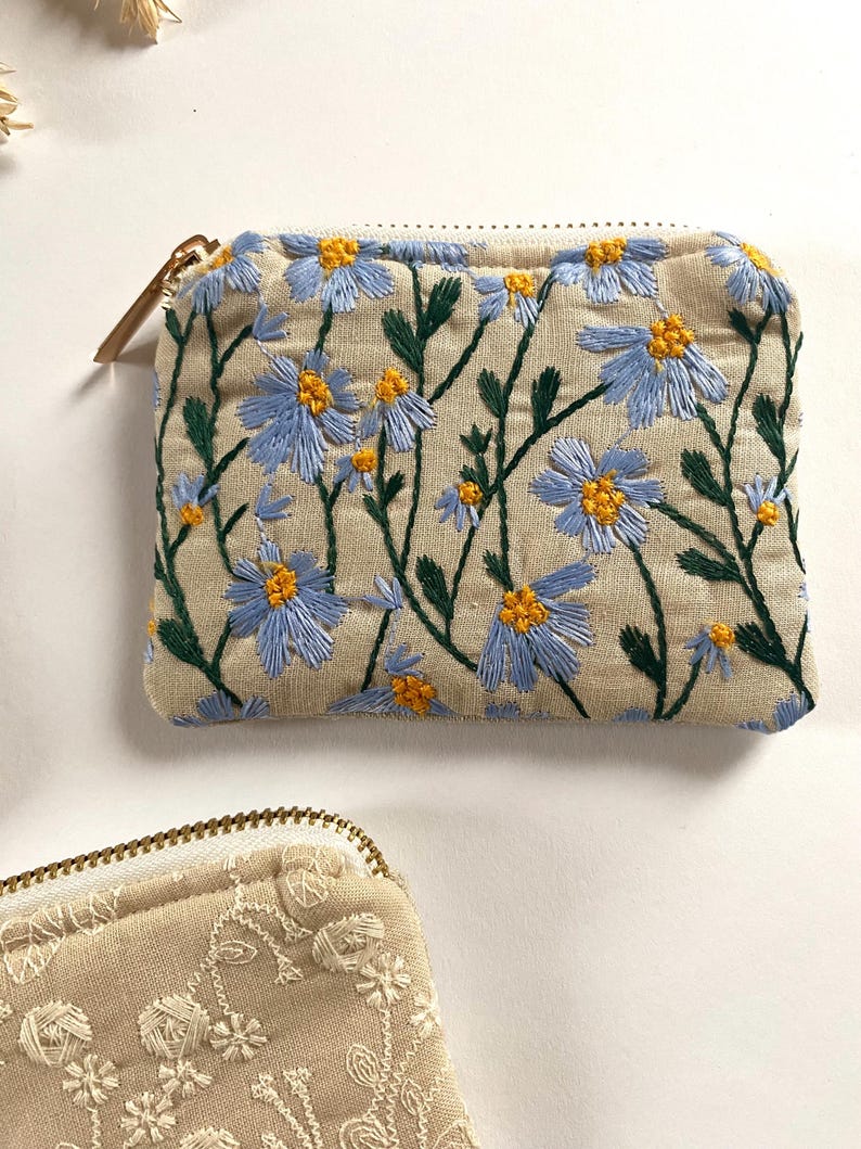 K&ouml;nnte beinhalten: Eine beige Rei&szlig;verschlusstasche mit gestickten blauen und gelben Blumen und gr&uuml;nen Stielen. Das florale Design bedeckt die Vorderseite der Tasche. Ein goldfarbener Rei&szlig;verschluss und Zipper sind sichtbar. Eine weitere Tasche mit floralem Design ist teilweise sichtbar.