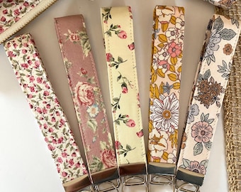 May include: Five fabric wristlet keychains featuring various floral patterns in shades of pink, yellow, cream, and muted green. Each strap is attached to a silver-toned metal hardware clamp with a circular split ring for keys.