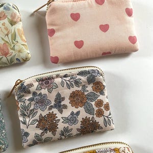 May include: Several small, zippered pouches with various floral and heart patterns. One pouch features a light pink background with repeating pink hearts. Other pouches display floral designs in shades of blue, brown, and yellow.