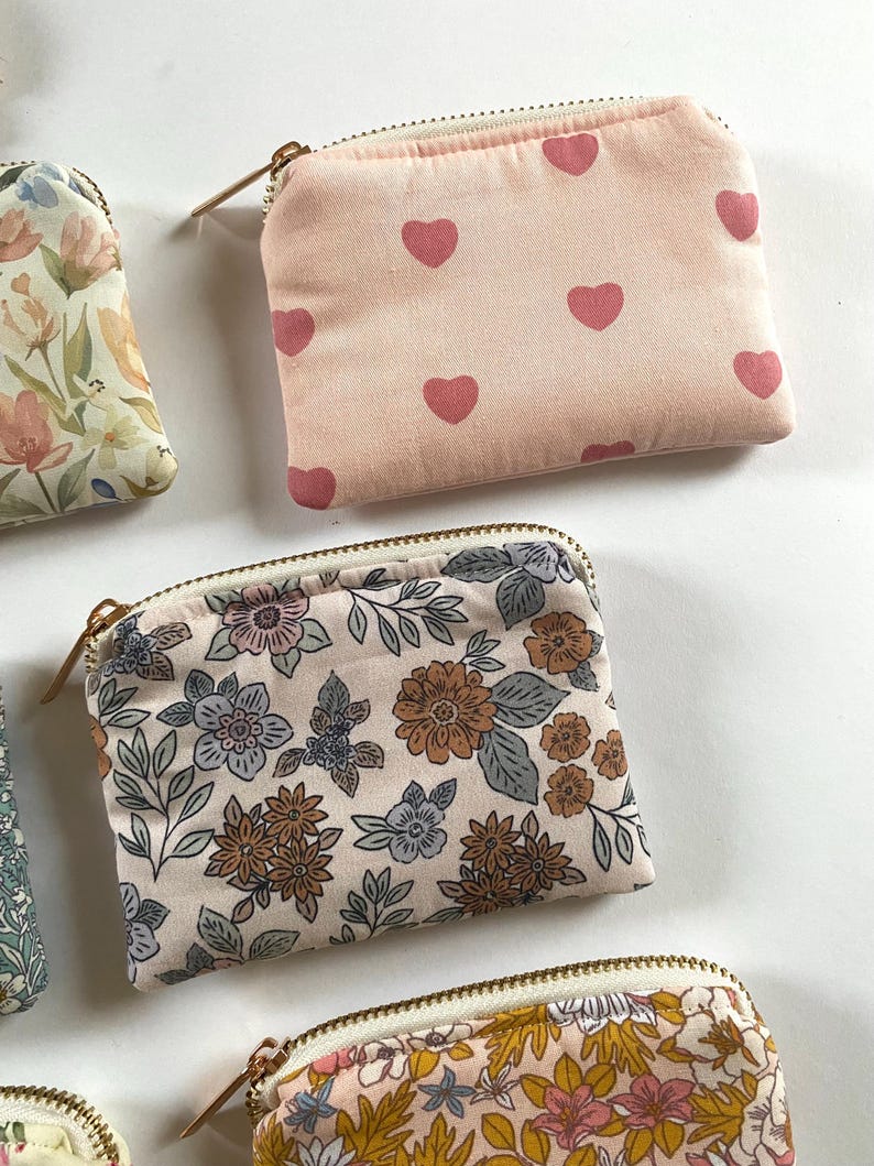 May include: Several small, zippered pouches with various floral and heart patterns. One pouch features a light pink background with repeating pink hearts. Other pouches display floral designs in shades of blue, brown, and yellow.
