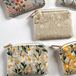 K&ouml;nnte beinhalten: Mehrere kleine, quadratische, bestickte M&uuml;nzb&ouml;rsen mit goldenen Rei&szlig;verschl&uuml;ssen. Die B&ouml;rsen weisen Blumenstickereien in verschiedenen Farben auf, darunter Rosa, Blau, Gelb und Wei&szlig;, auf beigem Hintergrund. Die Designs umfassen G&auml;nsebl&uuml;mchen, Rosen und andere Blumen.