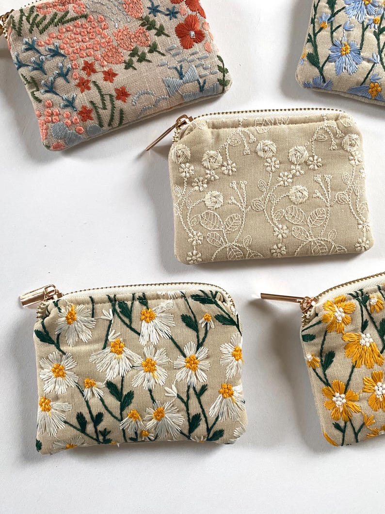 K&ouml;nnte beinhalten: Mehrere kleine, quadratische, bestickte M&uuml;nzb&ouml;rsen mit goldenen Rei&szlig;verschl&uuml;ssen. Die B&ouml;rsen weisen Blumenstickereien in verschiedenen Farben auf, darunter Rosa, Blau, Gelb und Wei&szlig;, auf beigem Hintergrund. Die Designs umfassen G&auml;nsebl&uuml;mchen, Rosen und andere Blumen.