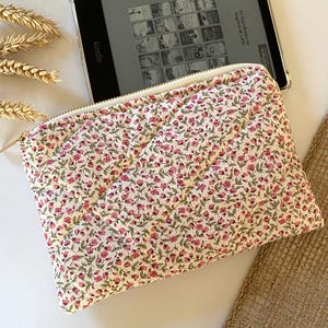 May include: A white quilted pouch with a zipper closure and a floral pattern featuring small pink flowers. The pouch is lying on a white surface next to a Kindle e-reader.
