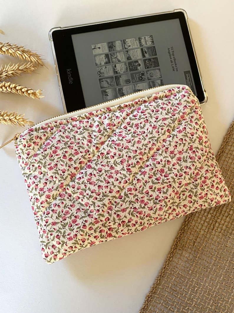 May include: A white quilted pouch with a zipper closure and a floral pattern featuring small pink flowers. The pouch is lying on a white surface next to a Kindle e-reader.