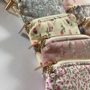 May include: A collection of small, floral-patterned pouches with gold-colored zippers. The pouches feature various pastel colors, including pink, blue, and cream, with delicate flower designs. The zippers are a prominent design element.