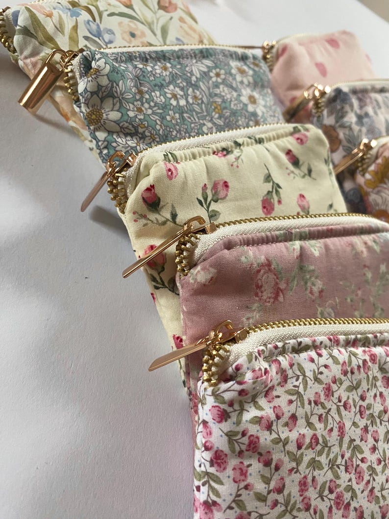 May include: A collection of small, floral-patterned pouches with gold-colored zippers. The pouches feature various pastel colors, including pink, blue, and cream, with delicate flower designs. The zippers are a prominent design element.