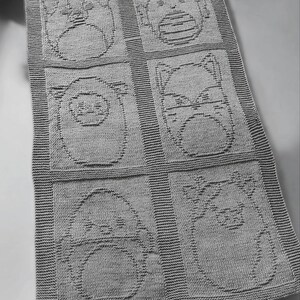 May include: A gray knitted baby blanket featuring six squares, each with a different animal face design. The animals include a bear, a bird, a cat, a pig, and two other creatures. The blanket has a ribbed border.