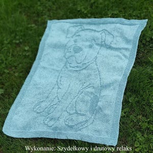 May include: A light blue knitted blanket with a detailed dog outline. The blanket has a textured appearance and a contrasting border. The text "Wykonanie: Szydełkowy i drutowy relaks" is visible at the bottom.