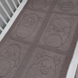 May include: A knitted baby blanket in a crib, featuring a repeating pattern of animal faces in a muted brown color. The blanket is rectangular and appears soft and cozy, perfect for a nursery.