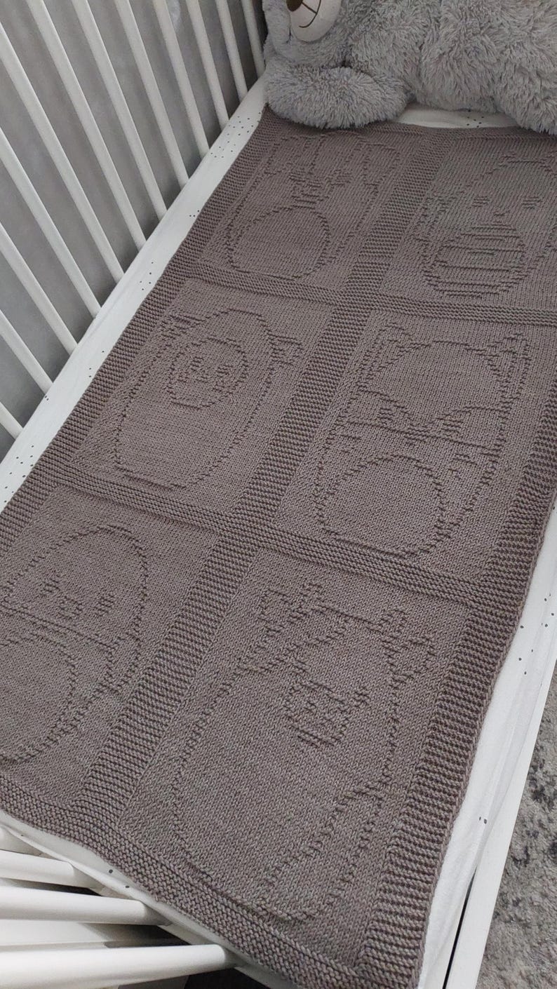 May include: A knitted baby blanket in a crib, featuring a repeating pattern of animal faces in a muted brown color. The blanket is rectangular and appears soft and cozy, perfect for a nursery.