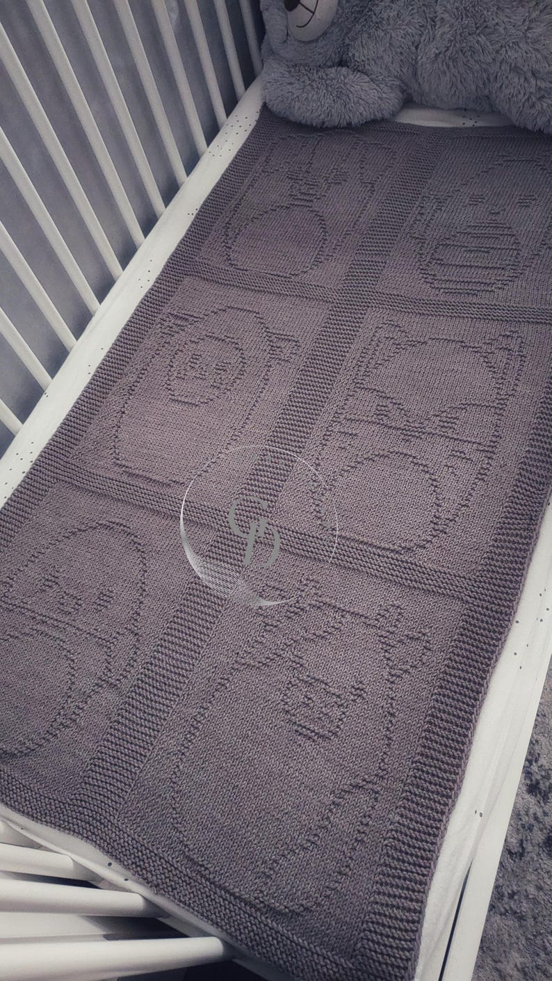 May include: A gray knitted baby blanket with a repeating pattern of cartoon-like images. The blanket is rectangular and appears to be made of soft yarn. The blanket is placed in a white crib with a gray stuffed animal.