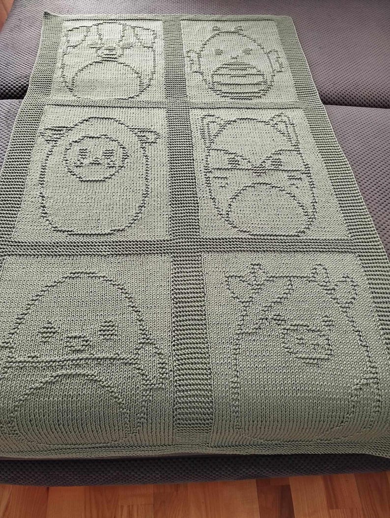 May include: A sage green knitted blanket featuring six squares, each with a different animal face design. The animals include a pig, a bear, a sheep, a fox, a penguin, and a cow. The blanket is made of soft material and is suitable for a baby or child.