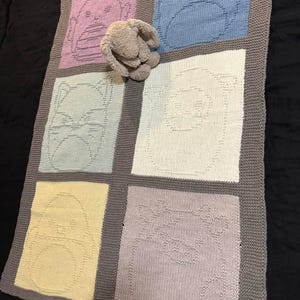 May include: A knitted baby blanket featuring six squares, each with a different animal face design in pastel colors. The blanket has a gray border and a stuffed bunny is resting on top.