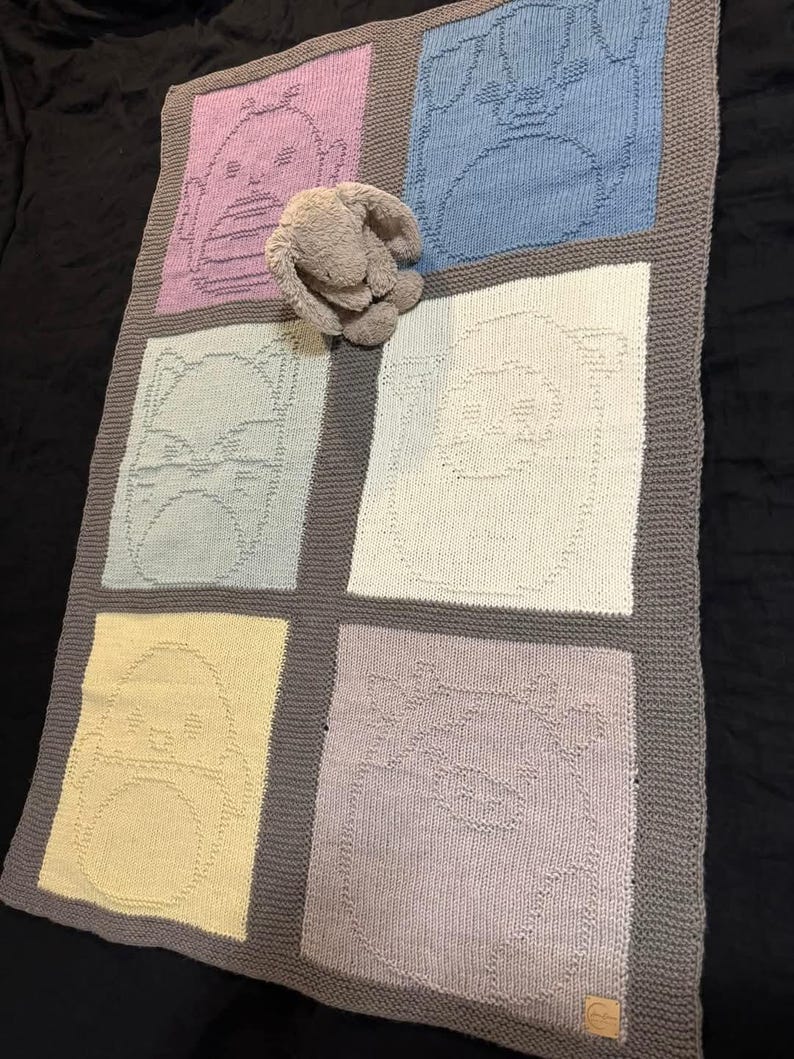 May include: A knitted baby blanket featuring six squares, each with a different animal face design in pastel colors. The blanket has a gray border and a stuffed bunny is resting on top.