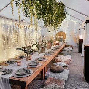 May include: A long wooden table with a white tablecloth and gray placemats is set for a dinner party. The table is decorated with string lights, greenery, and candles. There are pillows on the floor around the table, creating a cozy and intimate atmosphere.