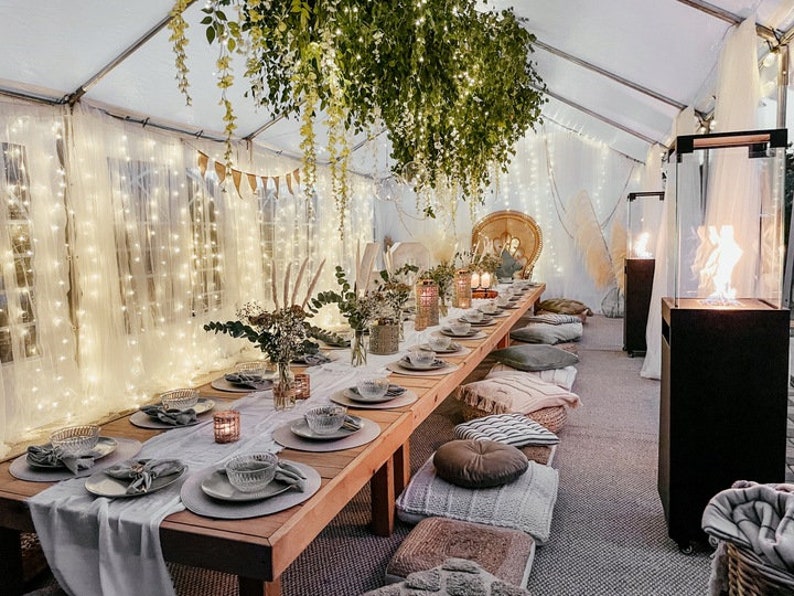 May include: A long wooden table with a white tablecloth and gray placemats is set for a dinner party. The table is decorated with string lights, greenery, and candles. There are pillows on the floor around the table, creating a cozy and intimate atmosphere.