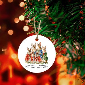 May include: A white ceramic Christmas ornament featuring a group of five bunnies, two adults and three children, in a line. The bunnies are wearing colorful clothing and the ornament has the text "Adult 1 name Child 1 Child 2 Adult 2 name Child 3 Child 4".