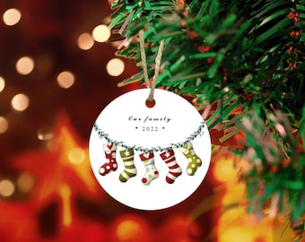 May include: White round Christmas ornament with a green garland and five colorful stockings hanging from it. The ornament says "Our family * 2022 *"