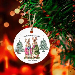 May include: White ceramic Christmas ornament with two bunnies wearing winter clothes. The ornament says "Our First Christmas Together" and has the names "Violet" and "Michael" at the bottom.