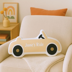 May include: A car-shaped pillow in a soft yellow color with black wheels and a white outline. The text "Jane's Ride" is printed on the side. The pillow is placed on a cream-colored chair.