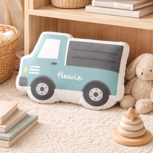 May include: A light blue truck-shaped pillow with the name "Howie" printed on the side. The pillow has black wheels, a gray truck bed, and a yellow headlight. A stuffed bunny and wooden stacking toy are also in the image.