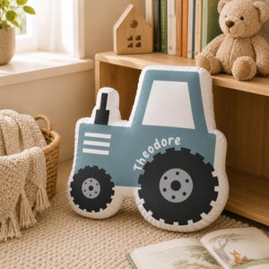 May include: A blue and white tractor-shaped throw pillow featuring a large black wheel and a smaller black wheel. The side of the tractor is printed with the name Theodore in white script lettering. The pillow has a white cabin window area.
